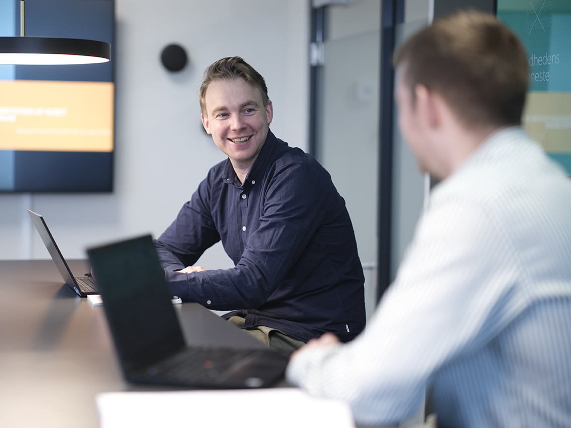 Mand sidder ved et mødebord og ser på en anden person, der sidder med en computer